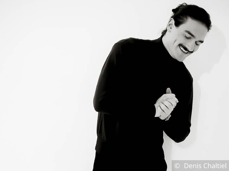 Photo en noir et blanc d'un jeune homme brun vêtu de noir qui pose les yeux fermé, souriant, les mains jointes.